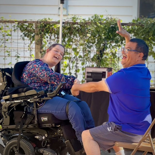 A woman in a power chair and a man seated in a folding chair each reach one arm toward the other. Both are smiling gleefully.