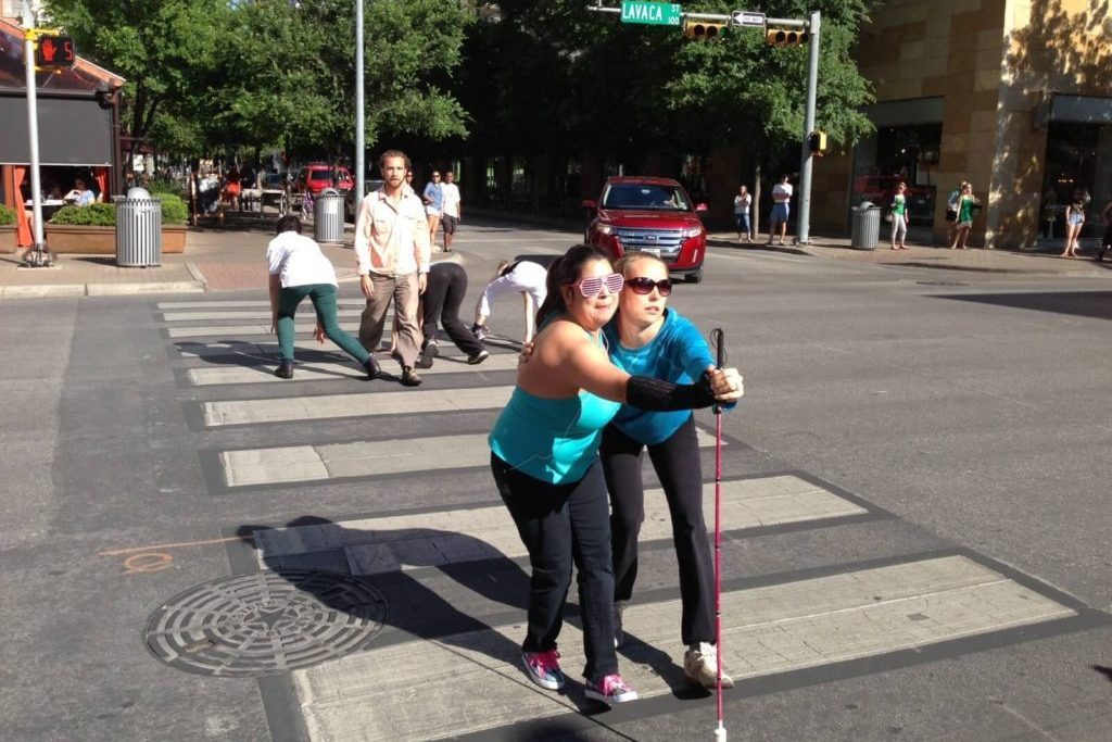 Two dancers, with and without disabilities, embrace each other and walk down a pedestrian crosswalk.