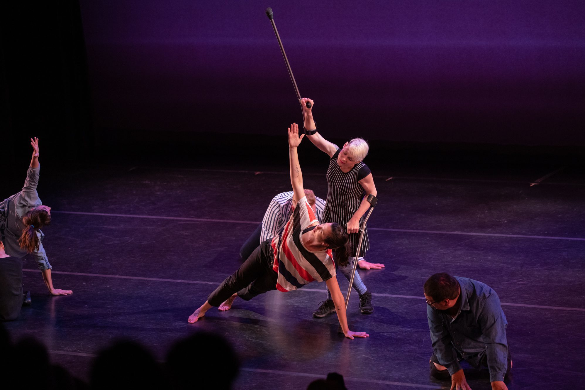 two dancers with and without disabilities perform on stage. One dancer is on the ground and the other stands behind them. Both dancers are reaching one arm towards the ceiling.