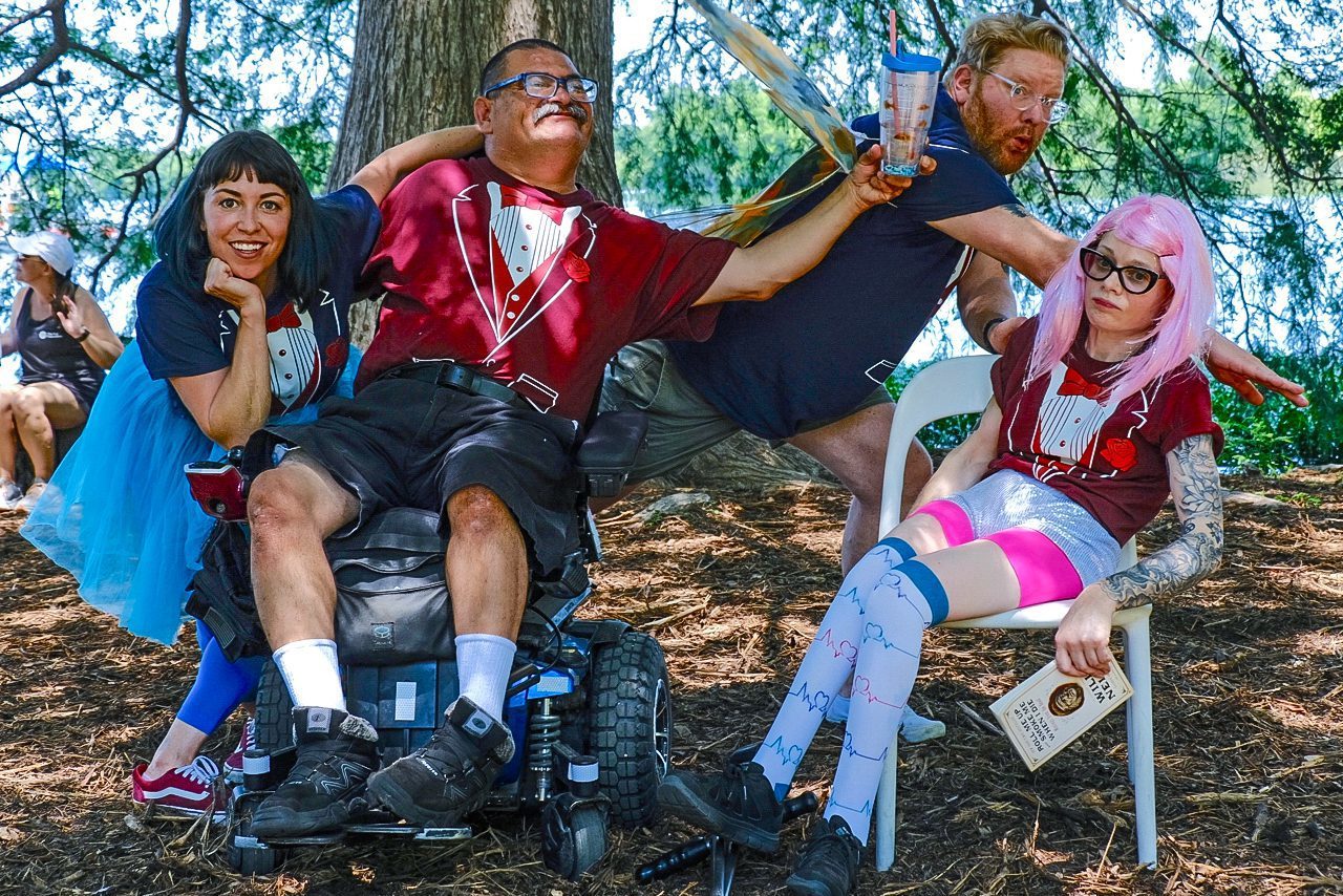 Dancers with and without disabilities wearing costumes, tutus, and wigs pose for a photo in a park.
