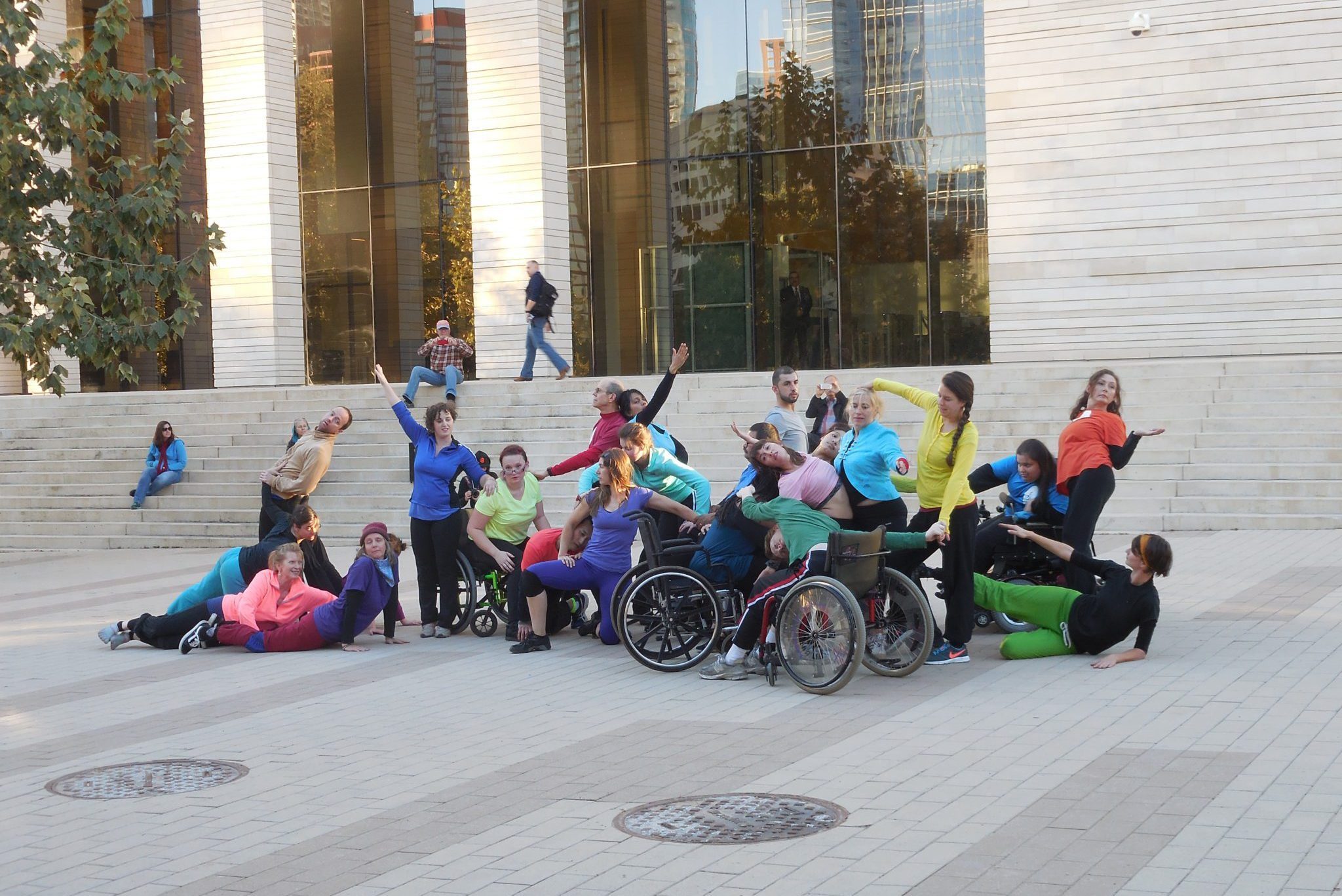 A group of dancers with and without disabilities perform outside in front of a large building. The dancers are in various poses and wearing brightly colored clothing.