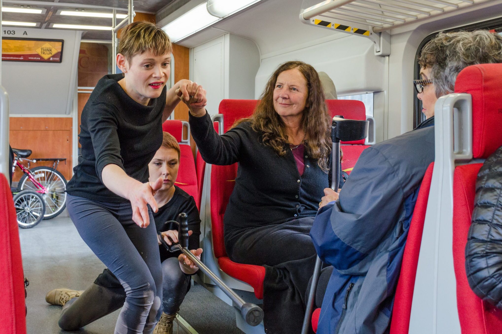 Tanya Winters and Silva Laukkanen dancing on a train. Tanya holds hands with a passenger as Silva passing Tanya's crutches to another passenger.