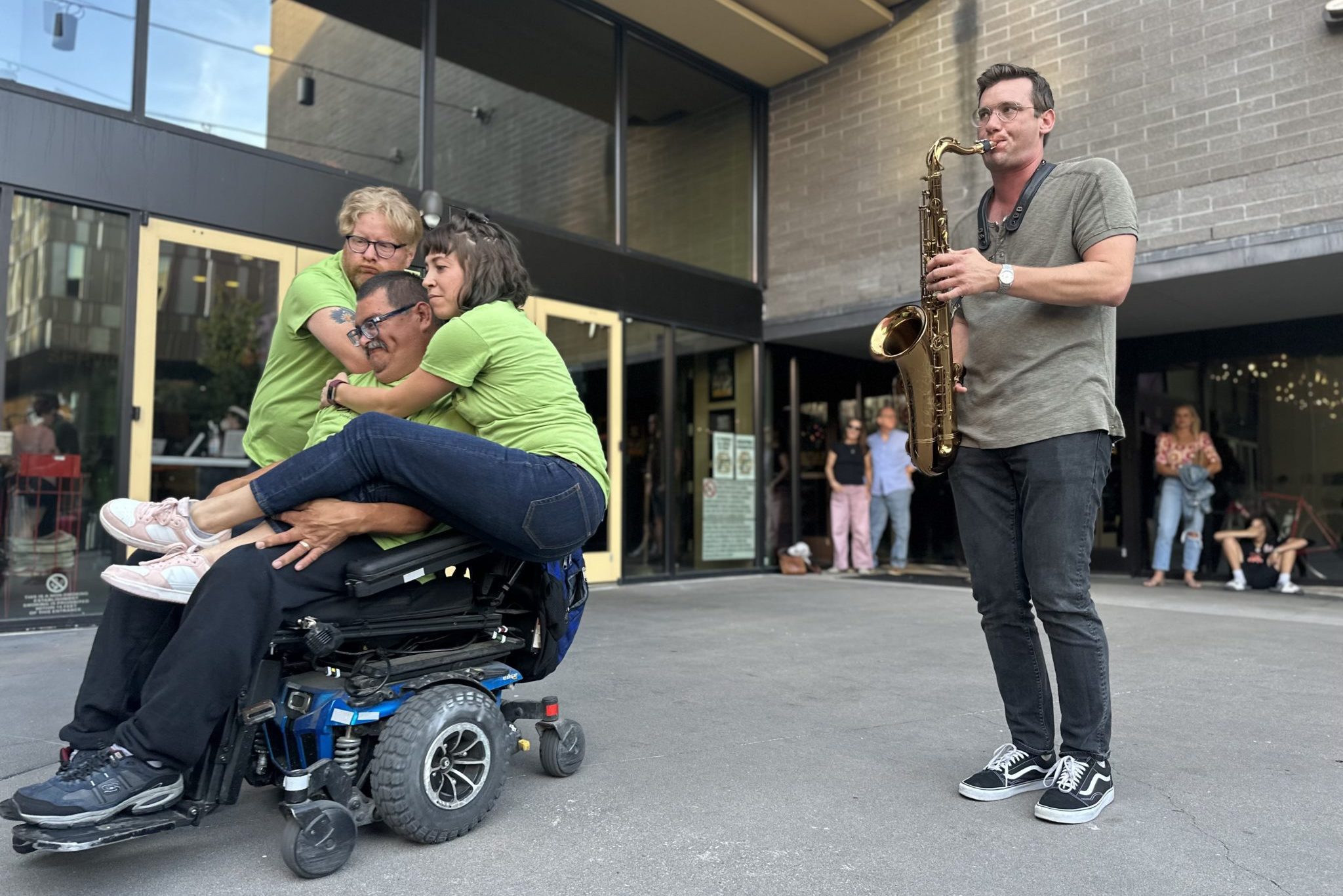 two stand-up dancers and one sit-down dancer perform with a person playing a saxophone outside. The two stand-up dancers hug the sit-down dancer and rest on their wheelchair.