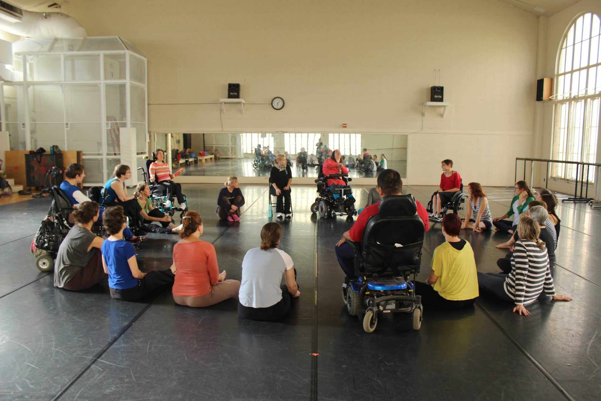 A group of dancers with and without disabilities sitting in a circle listening to someone speak.