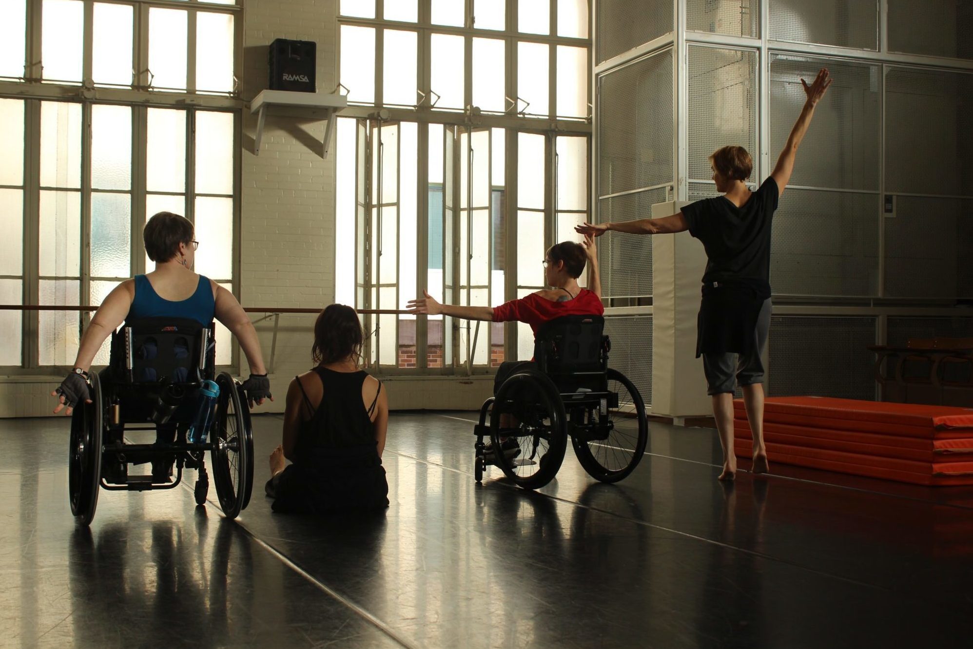 four dancers with and without disabilities positioned side by side face away from the camera. two dancers on the left have one arm reaching up and the other arm stretched to the side.