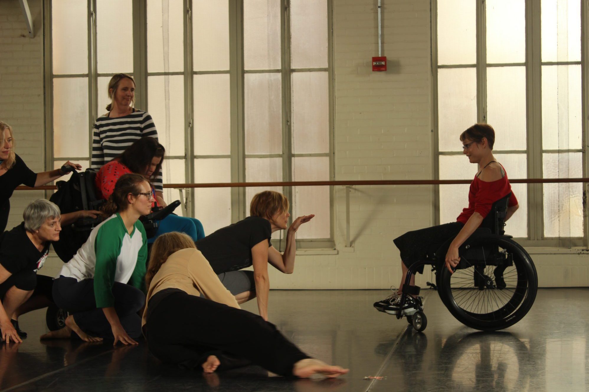 A group of 8 dancers with and without disabilities in a dance studio. 7 dancers crouch towards the ground smiling at a sit-down dancer who is facing them.