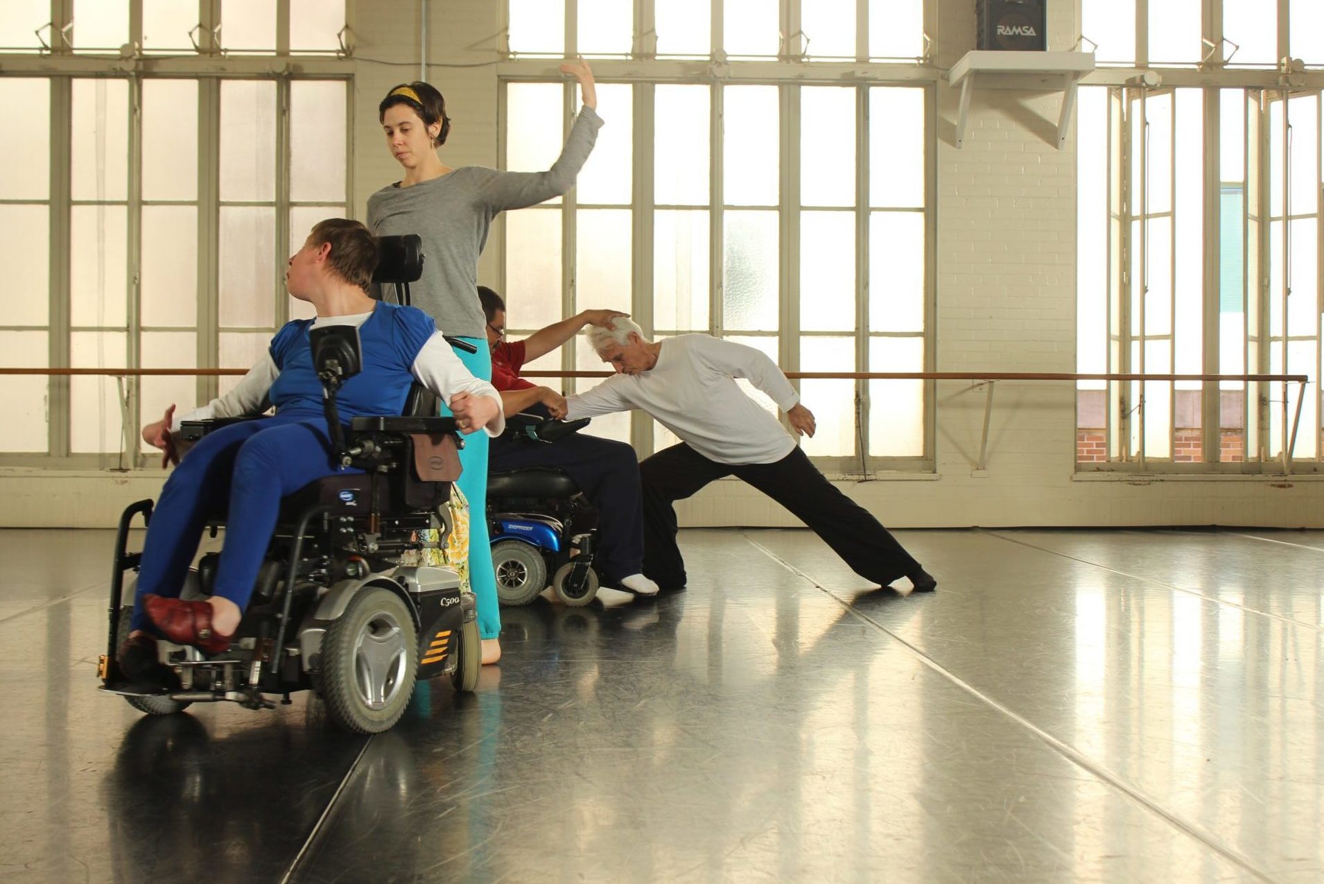 4 dancers with and without disabilities practice in a brightly lit dance studio with tall windows.