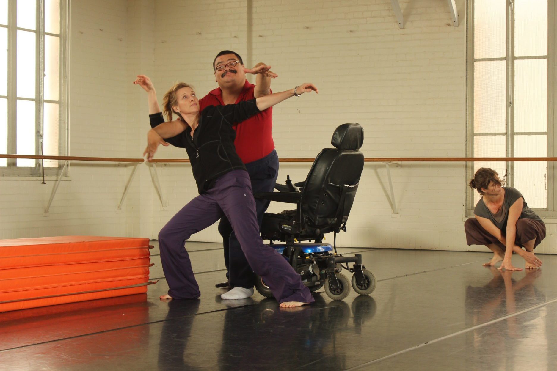 a wheelchair dancer and stand-up dancer practicing in a studio. The wheelchair dancer is standing embracing the stand-up dancer. Both are posed with one arm reaching out.