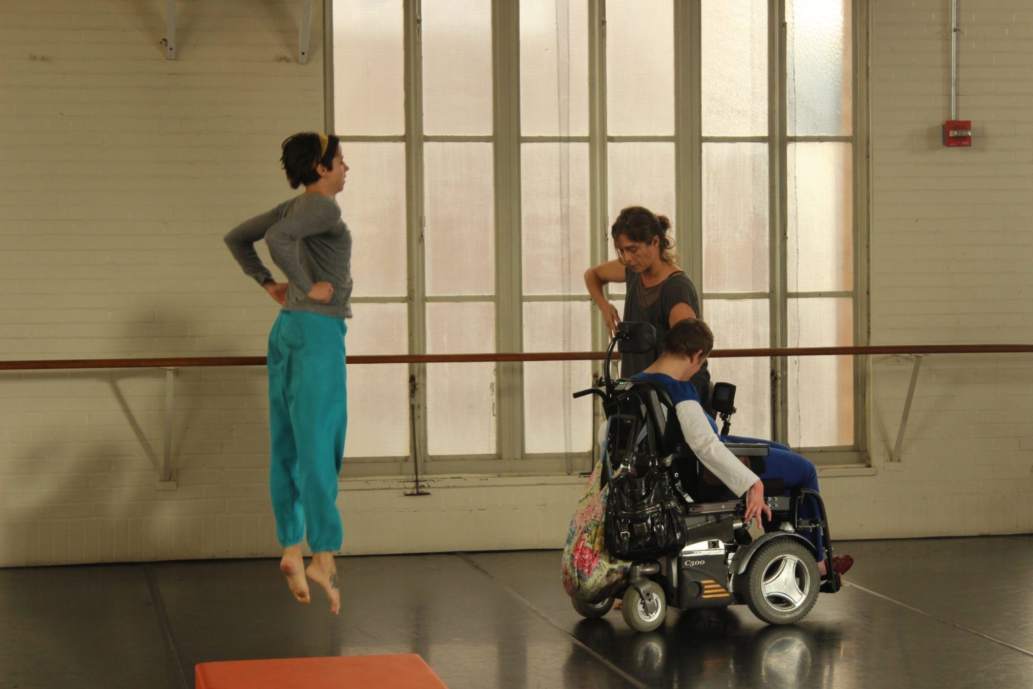 three dancers with and without disabilities practice in a studio space. One dancer is leaping into the air.