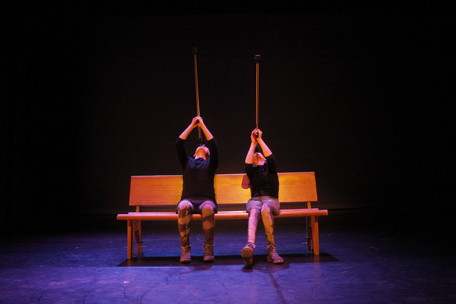 two performers with and without disabilities perform on stage. Both dancers are seated on a bench and are holding a walking crutch in the air.