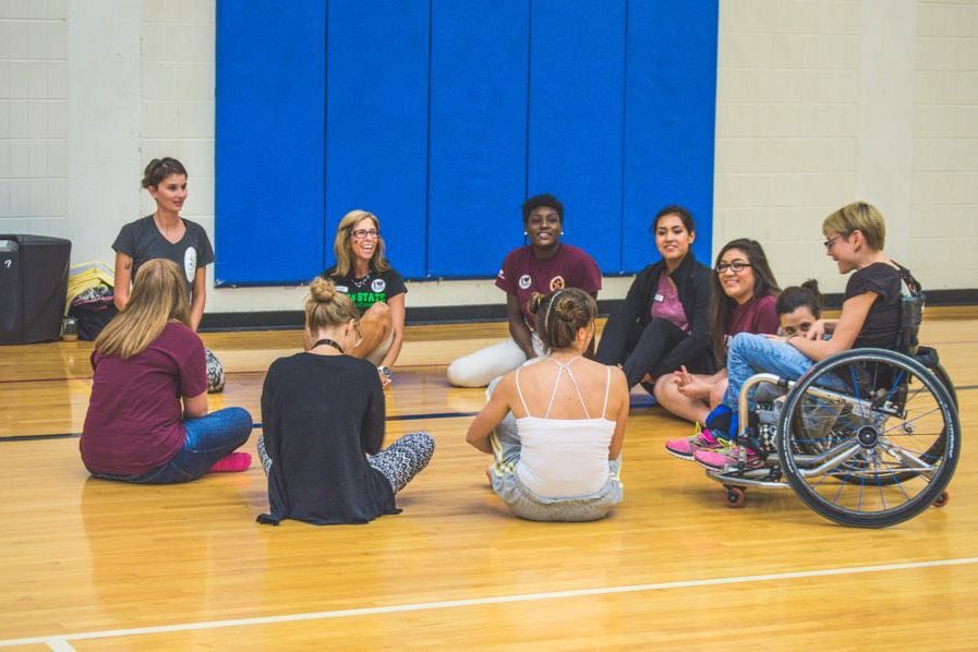A group of 11 dancers with and without disabilities sit in a circle in a student gymnasium listening to one person talk.