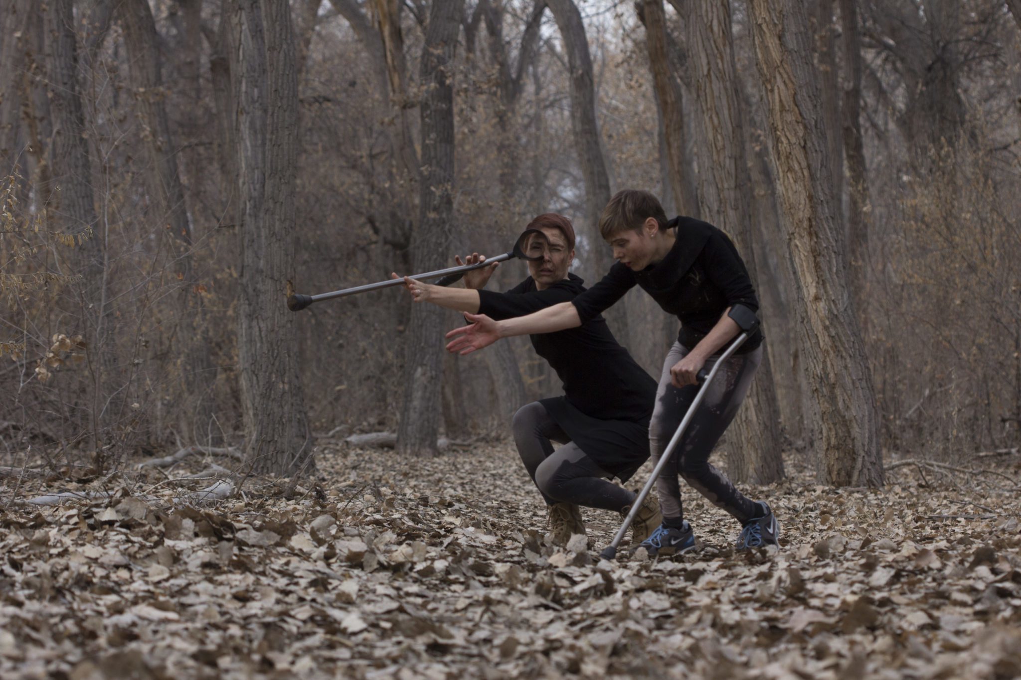 Silva Laukkanen and Tanya Winters performing in the woods. Silva is holding one of tanya's crutches and looks through it like a telescope.