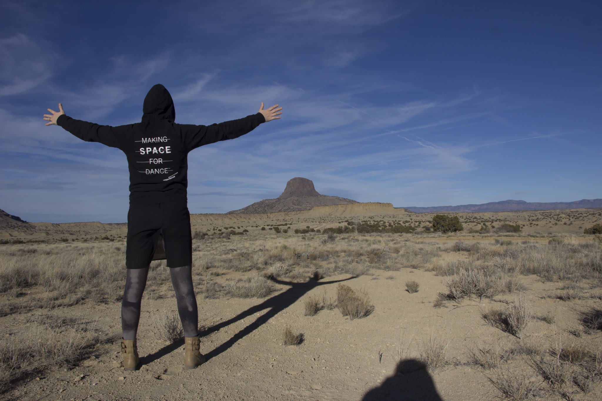 A dancer wearing a black hoodie poses in a desert landscape with a mountain in the distance.