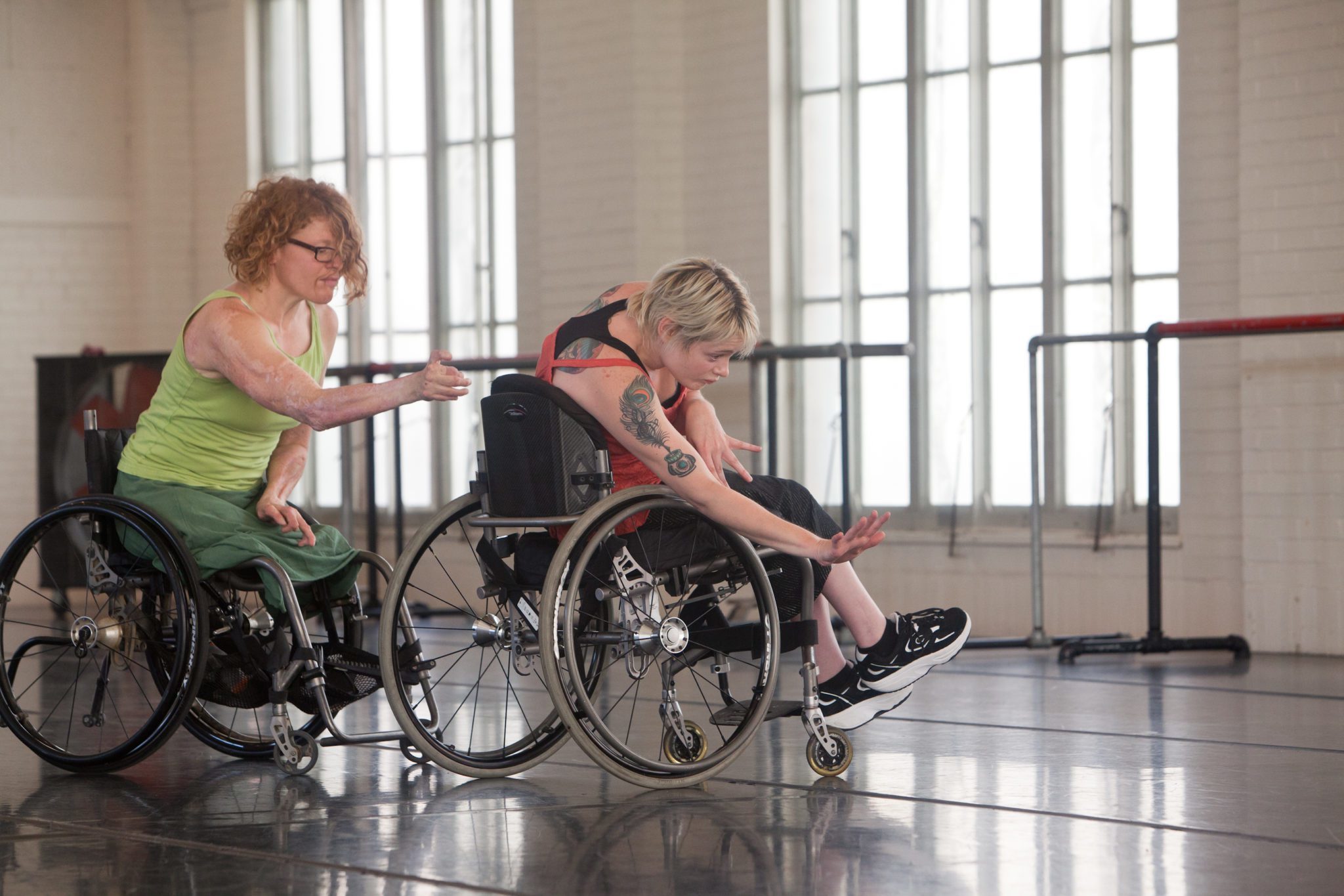 Two wheelchair dancers side by side with one arm reaching out in front of them.