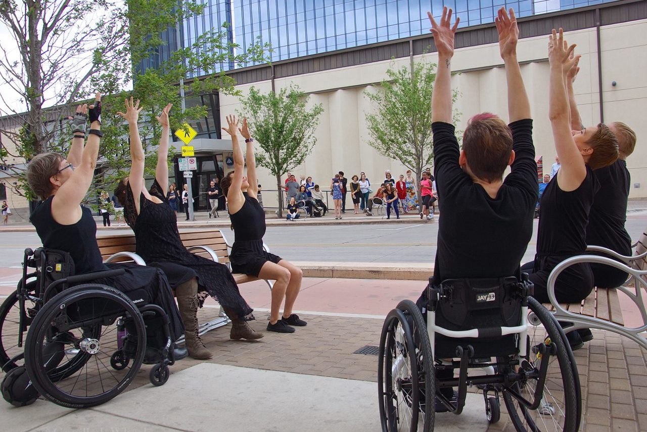 6 dancers with and without disabilities sit on outdoor benches with their arms stretch high.