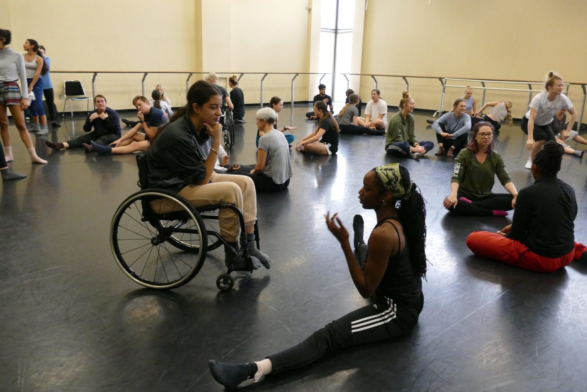 A large group of dancers with and without disabilities stretch in a studio space.