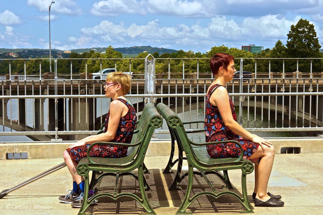 two performers with and without disabilities sit on park benches facing away from each other.